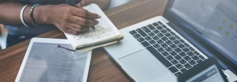 Person writing in notebook next to a laptop and tablet.